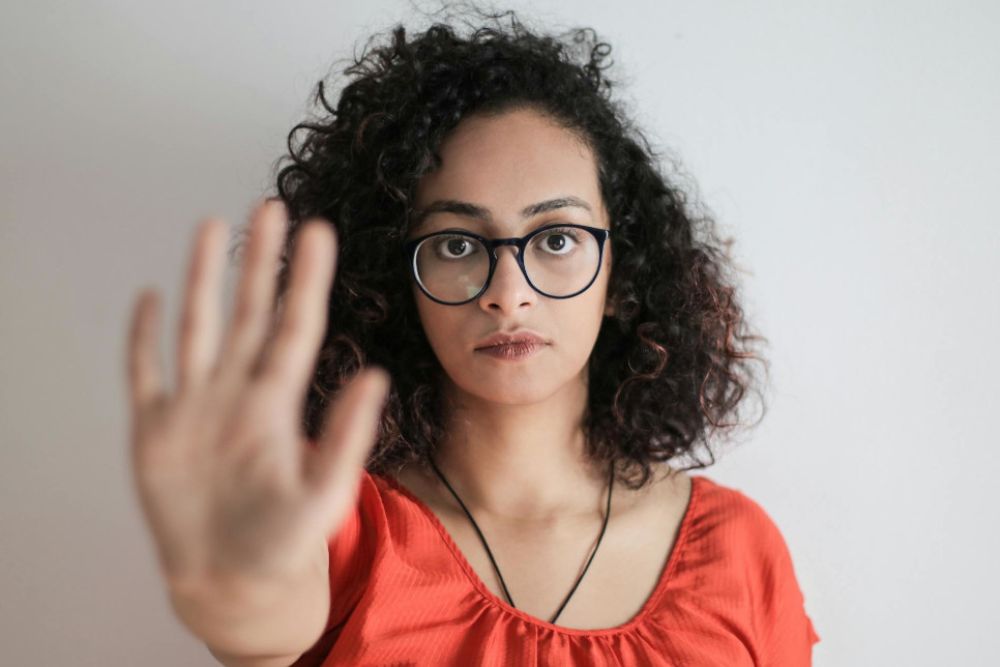 Eine Frau mit lockigen Haaren und Brille trägt ein rotes Oberteil. Sie schaut ernst in die Kamera und hebt ihre Hand nach vorne in einer Stop-Geste. Der Hintergrund ist neutral.