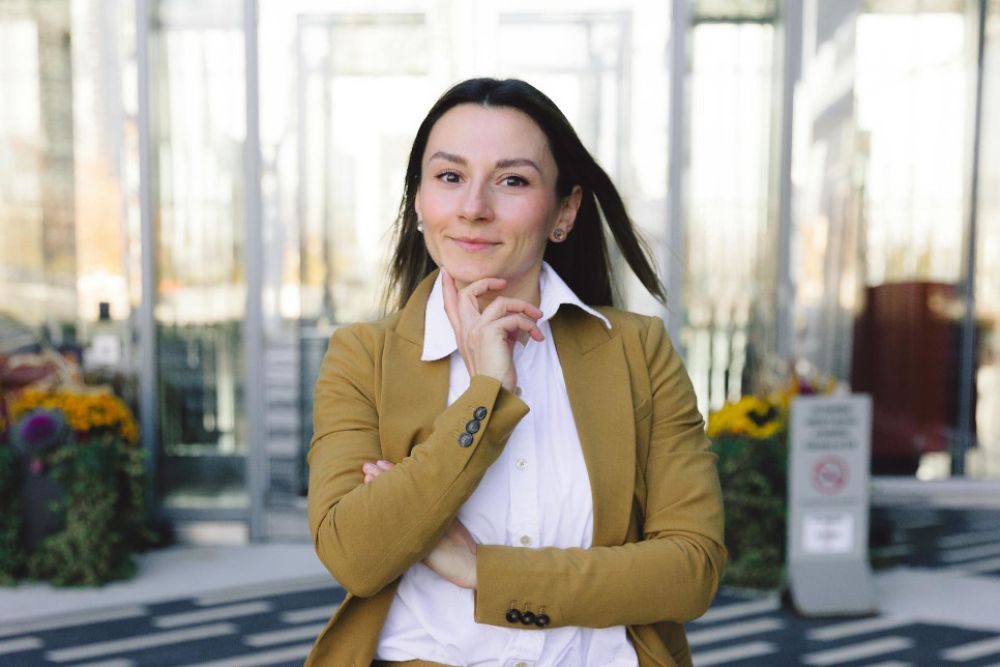 Eine junge Frau mit braunen Haaren trägt ein beigefarbenes Sakko und ein weißes Hemd. Sie steht draußen vor einem modernen Gebäude, lächelt leicht und stützt ihr Kinn mit der Hand. Der Hintergrund zeigt Glas und Pflanzen.