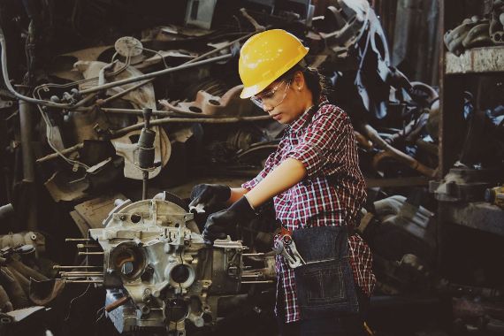 Das Bild zeigt eine Frau in einer Werkstatt, die konzentriert an einem metallischen Maschinenteil arbeitet. Sie trägt einen gelben Schutzhelm, eine Schutzbrille, schwarze Arbeitshandschuhe und ein rot-schwarz kariertes Hemd. Um ihre Hüfte befindet sich eine Werkzeuggürteltasche, die mit mehreren Werkzeugen bestückt ist.