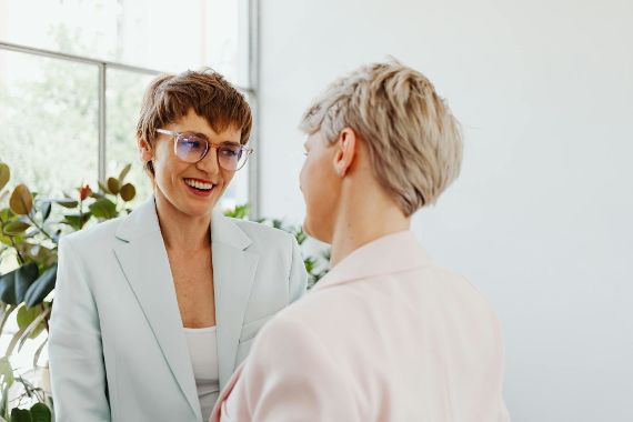 Zwei Frauen stehen sich gegenüber. Die linke Frau hat einen Kurzhaarschnitt, eine Brille und einen hellblauen Blazer. Die rechte Frau ebenfalls einen Kurzhaarschnitt und einen hellrosa Blazer an.